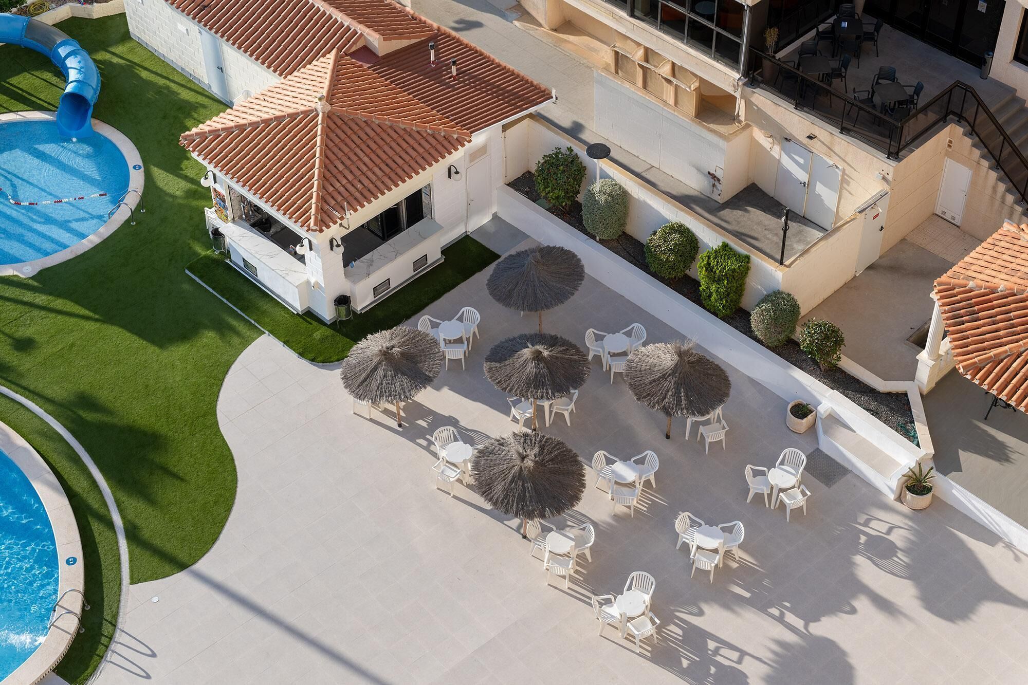 An aerial view of a resort with a swimming pool, outdoor seating area with umbrellas, and a building with a tiled roof.