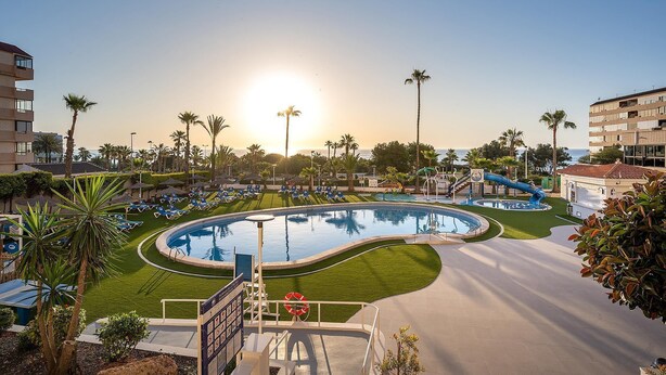 A resort pool area with a circular pool, sun loungers, palm trees, and a clear view of the ocean.