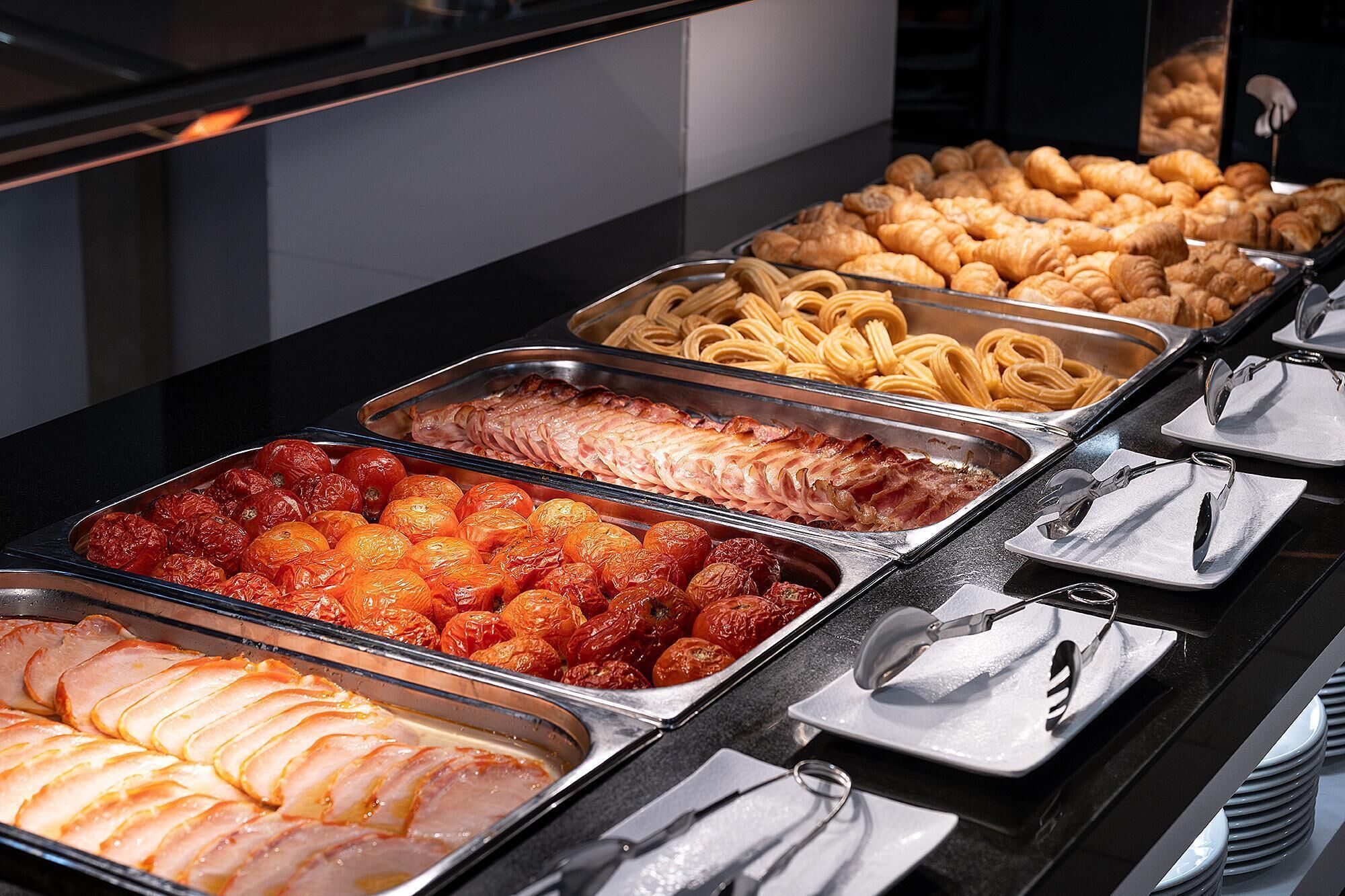 A buffet with various food items including sliced ham, roasted tomatoes, pasta, and pastries.