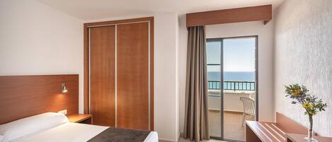 Quarto de hotel com cama, escrivaninha, cadeira e varanda com vista para o mar.