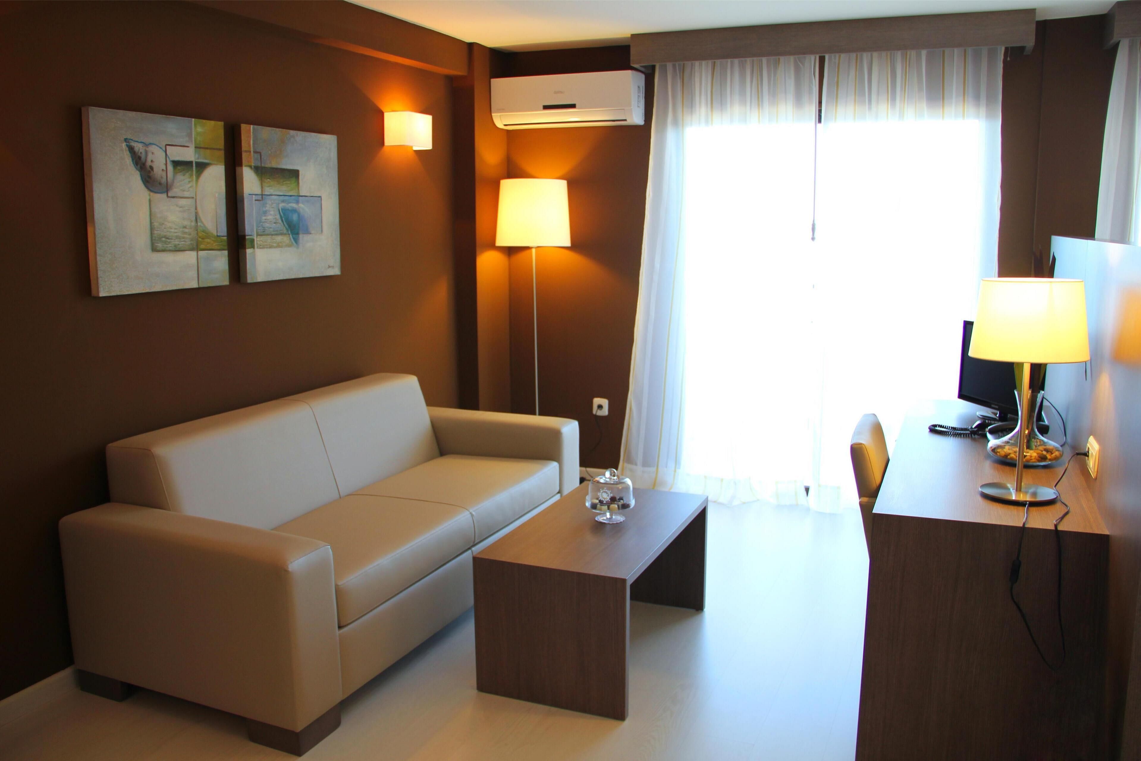 Een moderne hotelkamer met een bank, een houten bijzettafeltje, een bureau met een lamp en twee wandgemonteerde airconditioners.