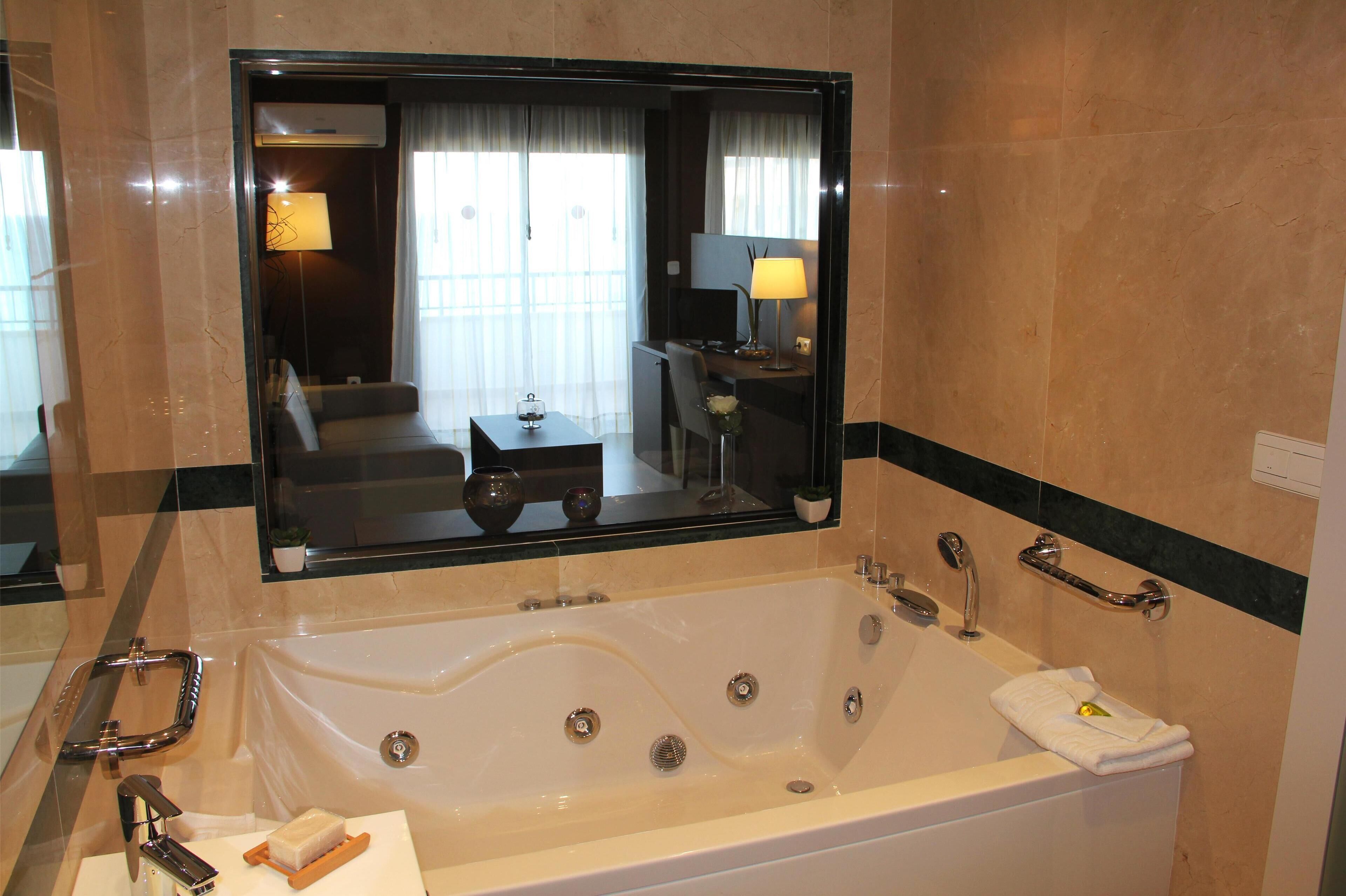 A modern bathroom with a large bathtub, a mirror, and a seating area visible in the reflection.
