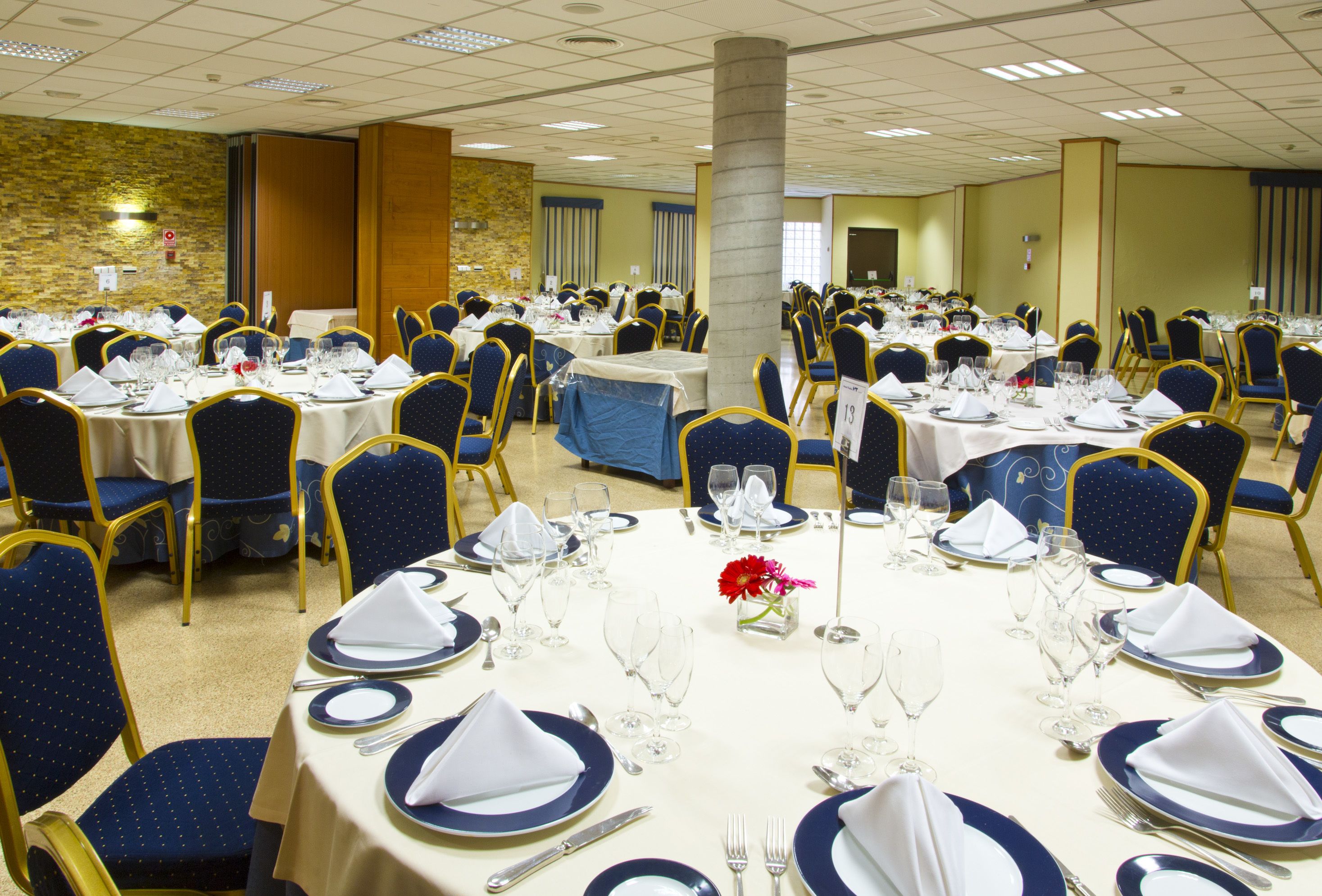 Un salón de banquetes con mesas redondas dispuestas para cenar, con manteles azules y blancos, sillas doradas y un pilar central.