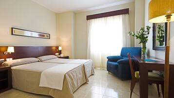 A hotel room with two beds, a desk with a chair, a lamp, and a large window with curtains.