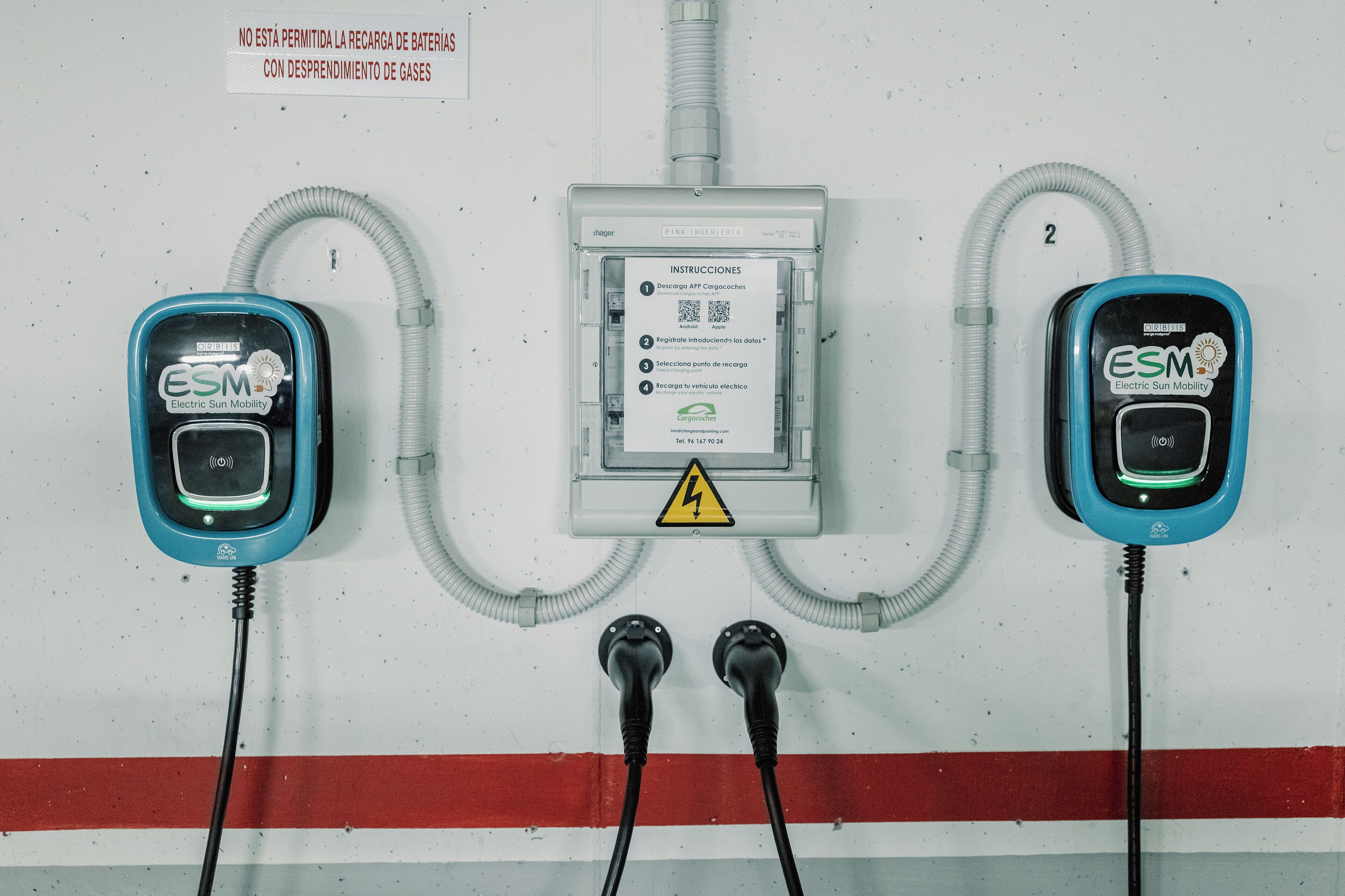 Two electric vehicle charging stations with cables connected to a wall-mounted box, and a warning sign above the box.