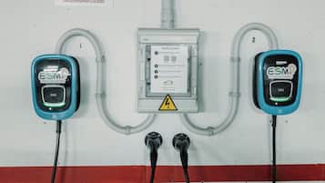 Dos estaciones de carga para vehículos eléctricos con cables conectados a una caja montada en la pared, y un cartel de advertencia sobre la caja.