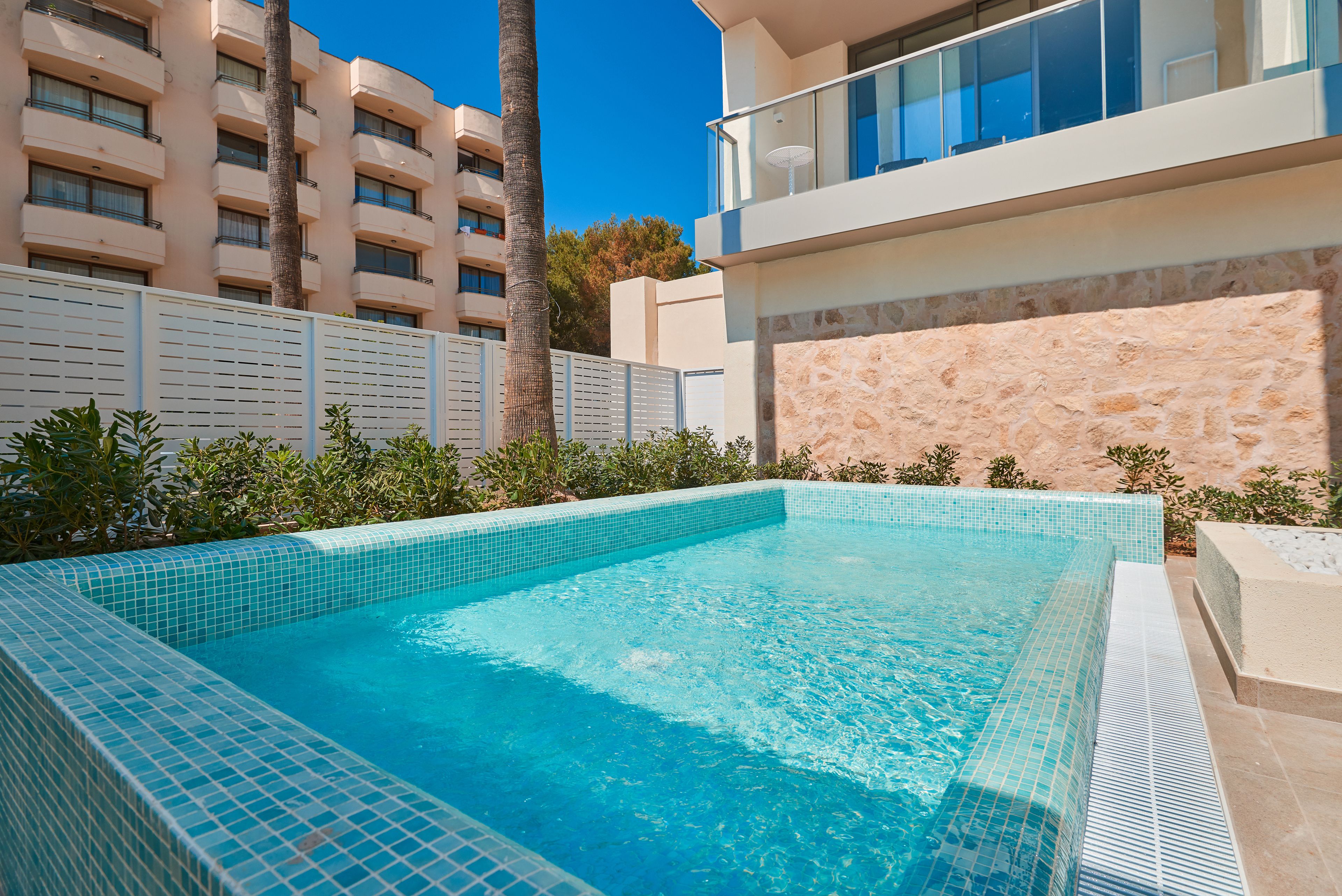 Una piscina con bordes de azulejos, rodeada por una valla blanca, palmeras y un edificio con balcones.