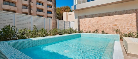Una piscina con bordes de azulejos, rodeada por una valla blanca, palmeras y un edificio con balcones.