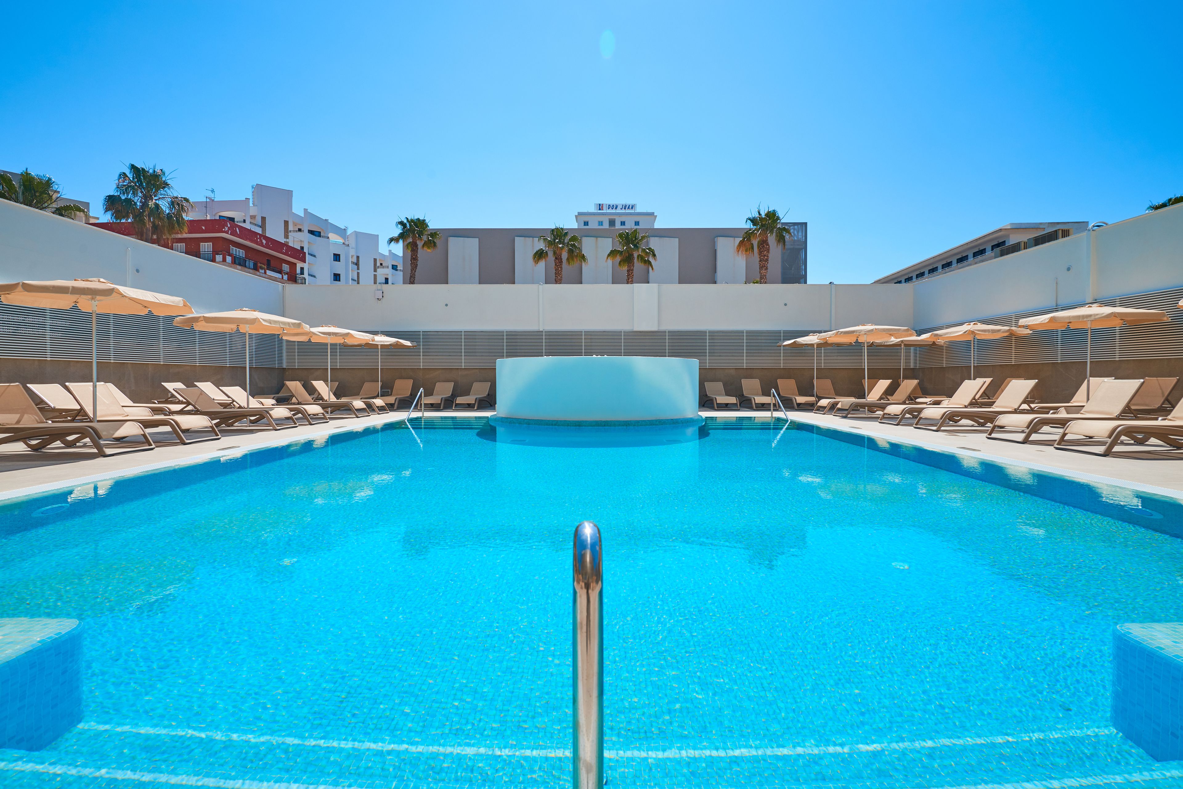 Piscina con sillas de descanso y sombrillas, rodeada de edificios.
