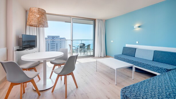 A modern hotel room with a balcony, a sofa, a dining table, and chairs. - Protur Atalaya Apartamentos (Son Servera)