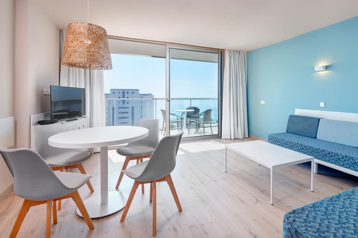 A modern hotel room with a balcony, a sofa, a dining table, and chairs.