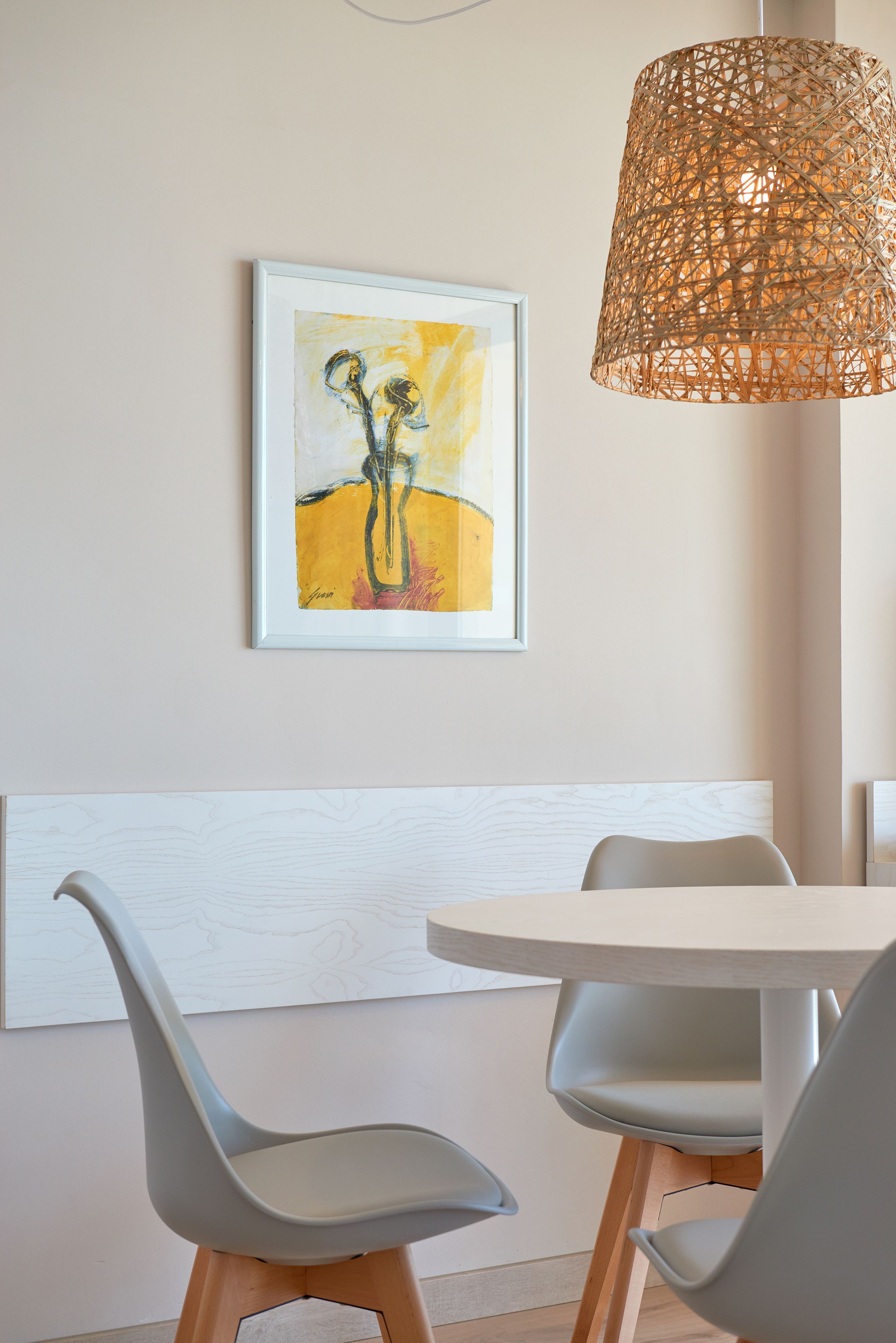 A modern dining area with a round table, two grey chairs, a woven pendant light, and a framed abstract painting on the wall.