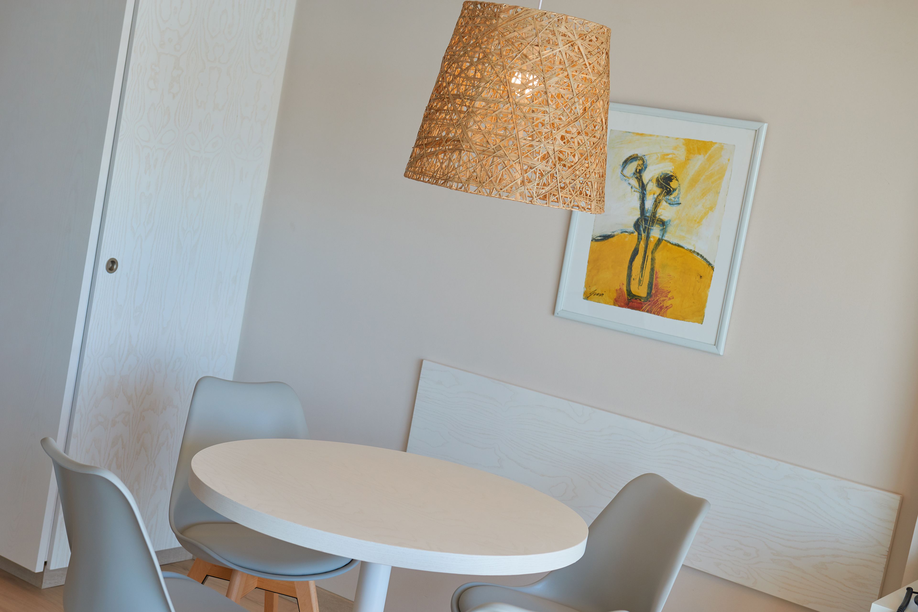 A dining area with a round table and four chairs, a woven pendant light, and a framed artwork on the wall.