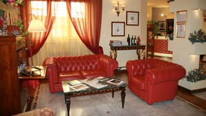 Una sala de estar con sofás de cuero rojo, una mesa de centro con libros y una lámpara. Hay cortinas, cuadros enmarcados en la pared y un mostrador de recepción al fondo.