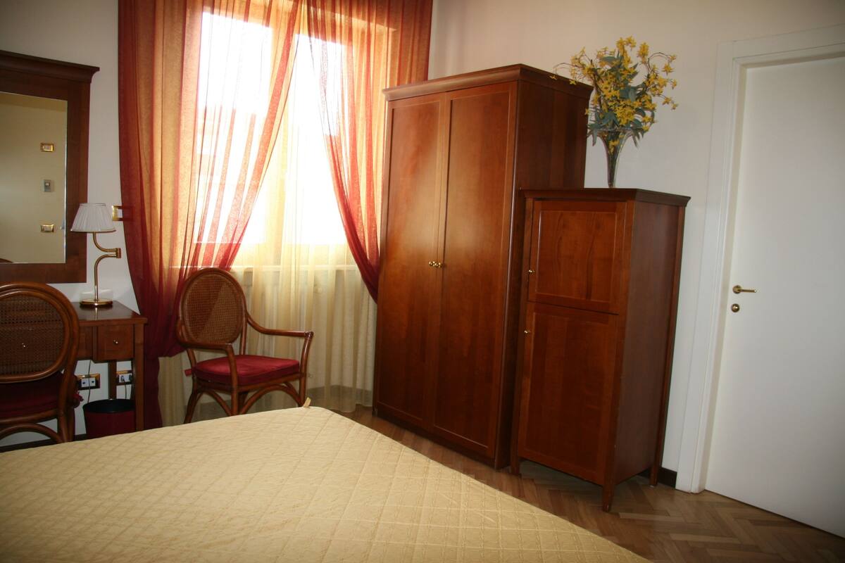 A bedroom with a bed, a wooden wardrobe, a chair, a desk, and a lamp.
