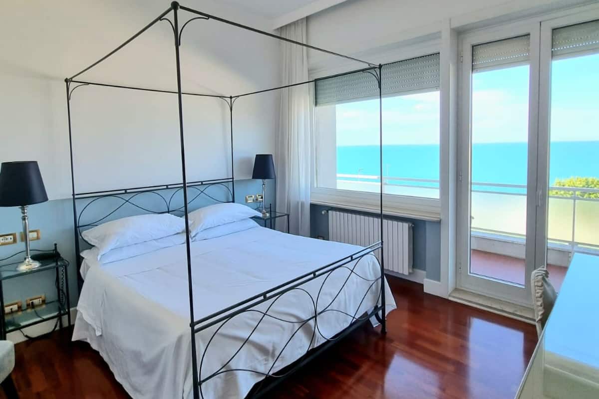 A bedroom with a four-poster bed, two bedside tables, a nightstand, and a view of the sea through large windows.