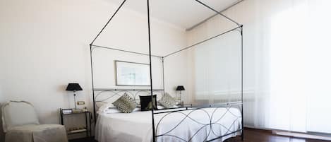 A four-poster bed with a white bedspread and decorative pillows, a bedside table with a lamp, and a chair.