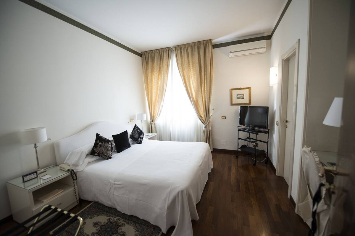 A hotel room with a bed, bedside table, lamp, TV, and a small desk.