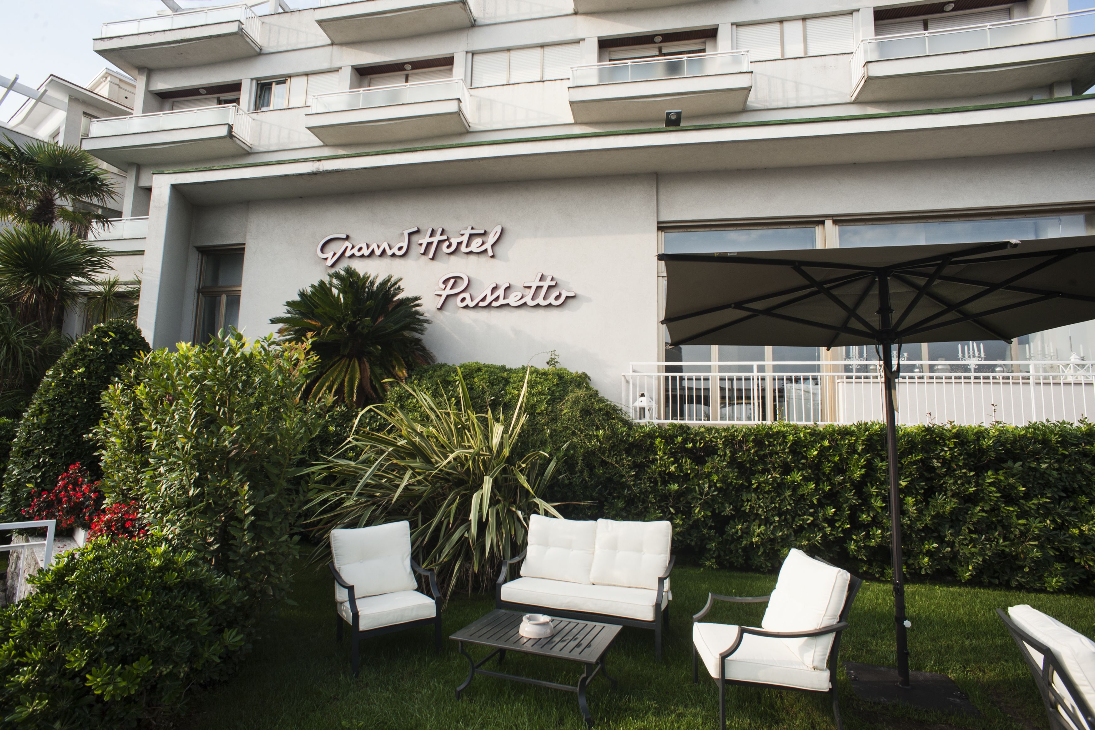 A hotel exterior with a sign reading "Grand Hotel Passetto," outdoor seating with white furniture, and a well-maintained garden.
