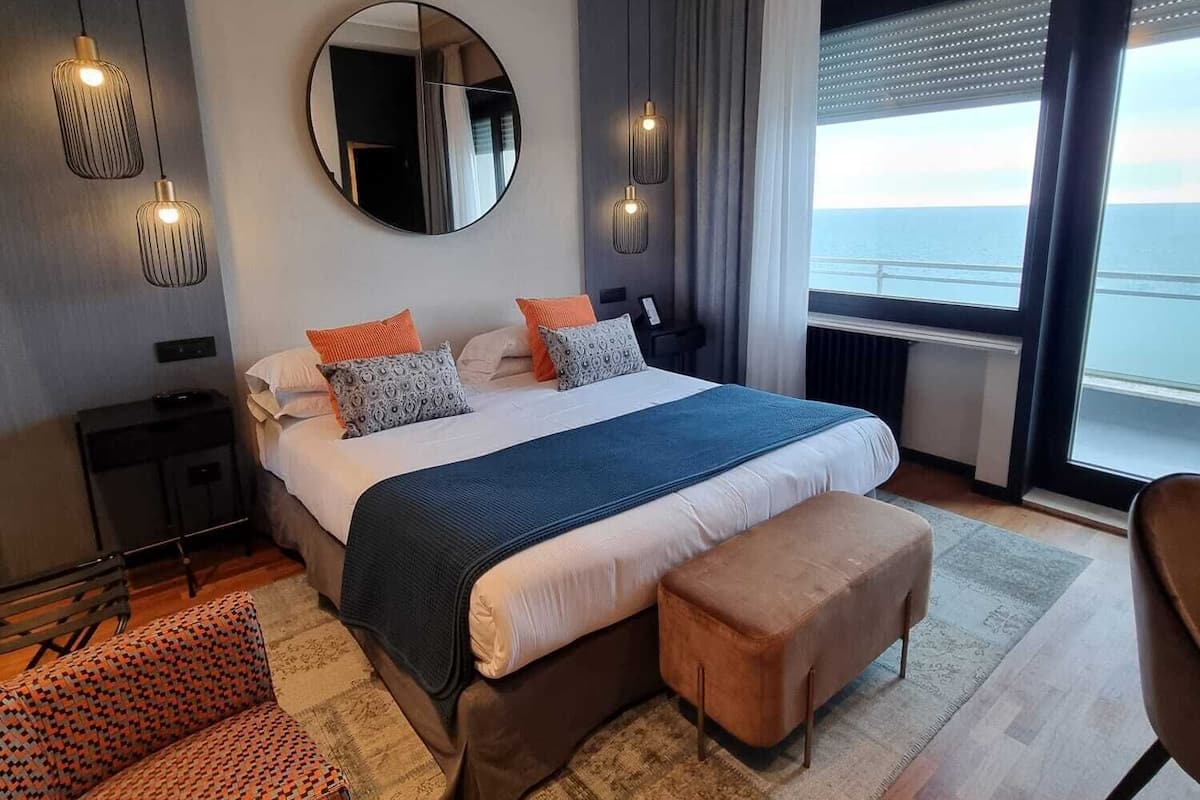 A modern bedroom with a large bed, a round mirror, pendant lights, a view of the sea, and a patterned rug.