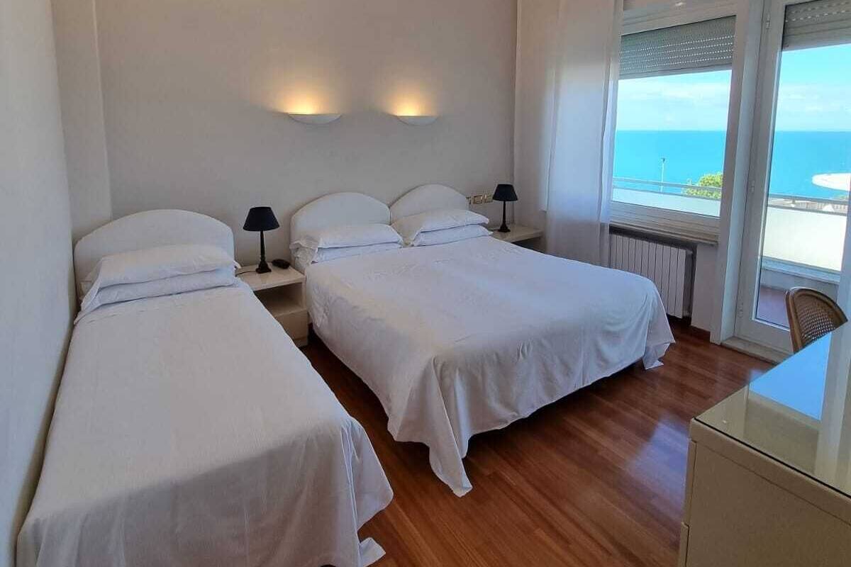 A hotel room with two single beds, a wooden floor, a window with a view of the sea, and a small desk with a chair.