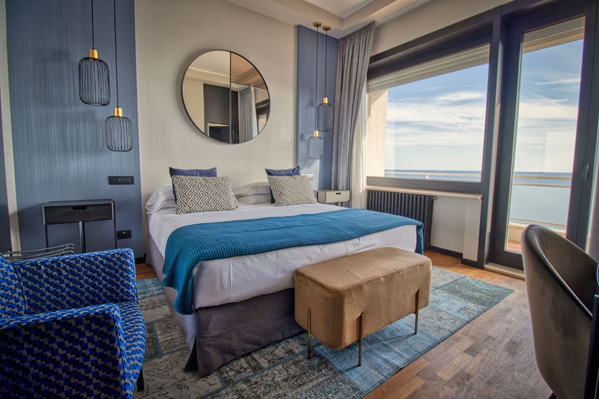 A modern hotel room with a large bed, a sitting area with a sofa and armchair, a small table, and a view of the sea through large windows.