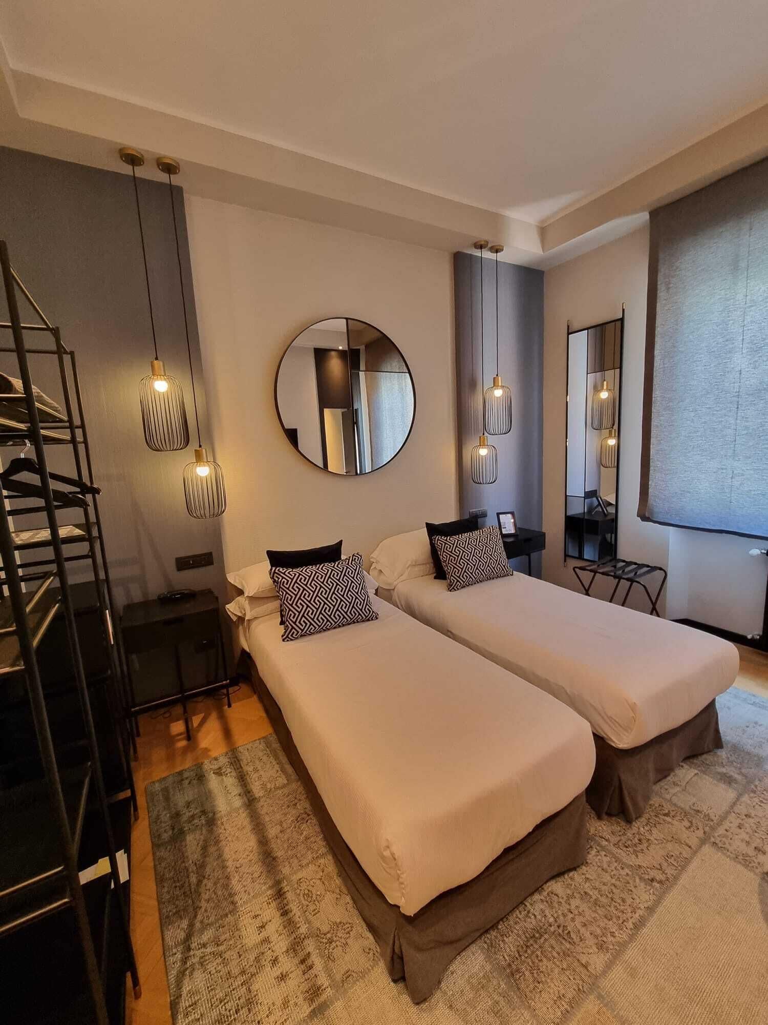 A modern bedroom with two beds, a round mirror, hanging pendant lights, and a staircase.