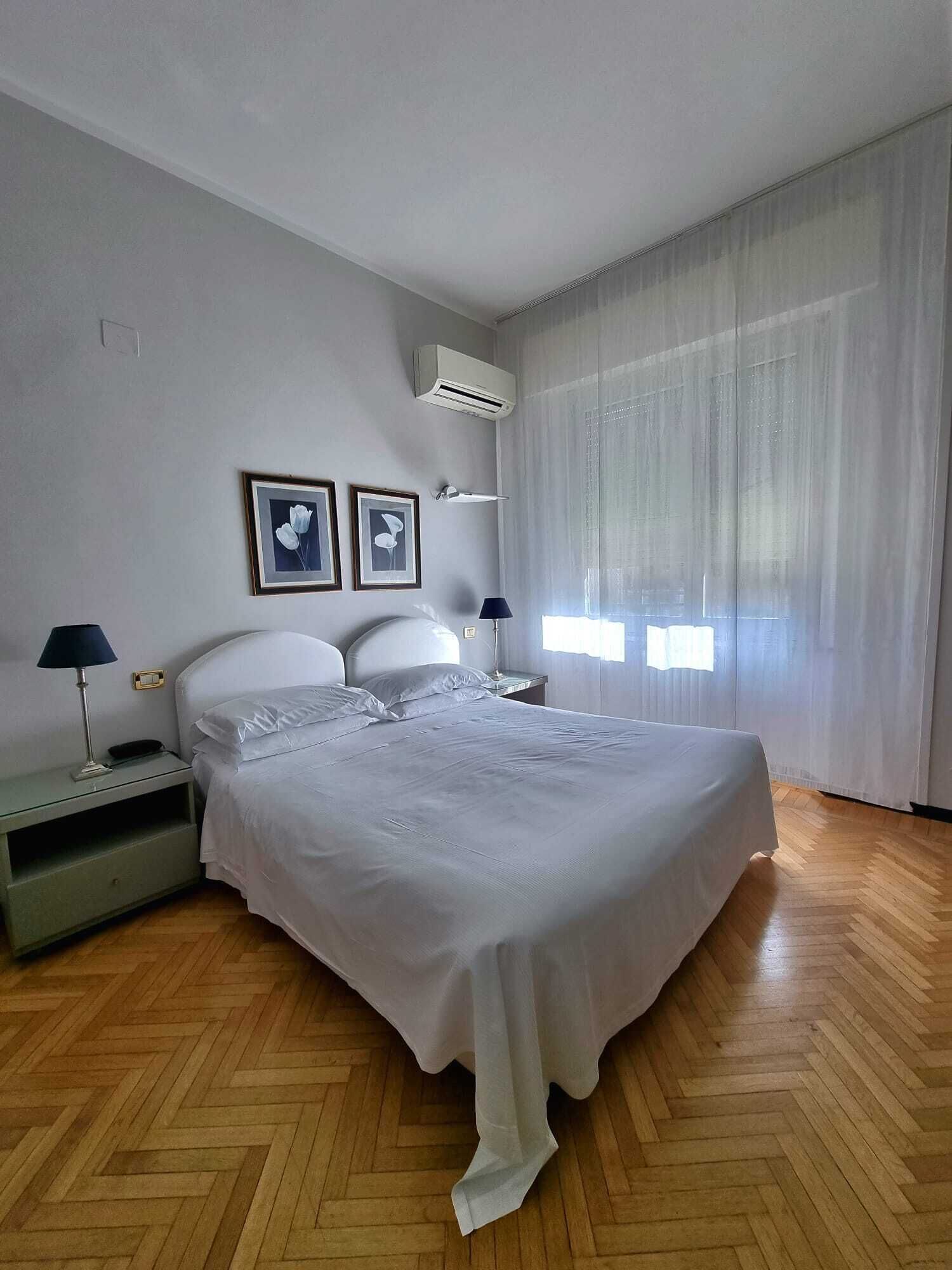 Un dormitorio con una cama grande, dos mesitas de noche con lámparas, un aire acondicionado de pared y dos cuadros enmarcados.