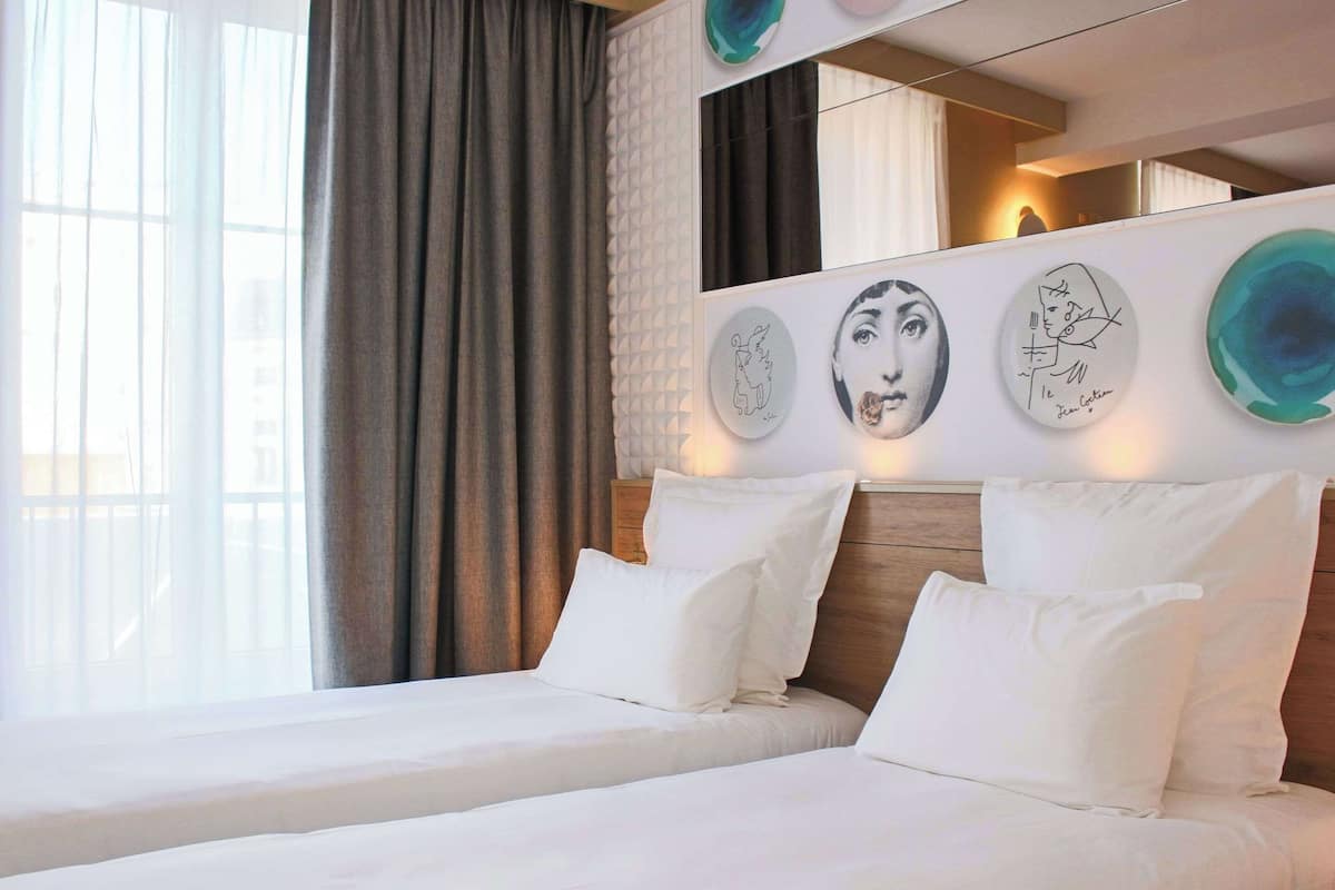 Une chambre d’hôtel avec deux lits, une grande fenêtre avec des rideaux et des œuvres d’art accrochées au mur.