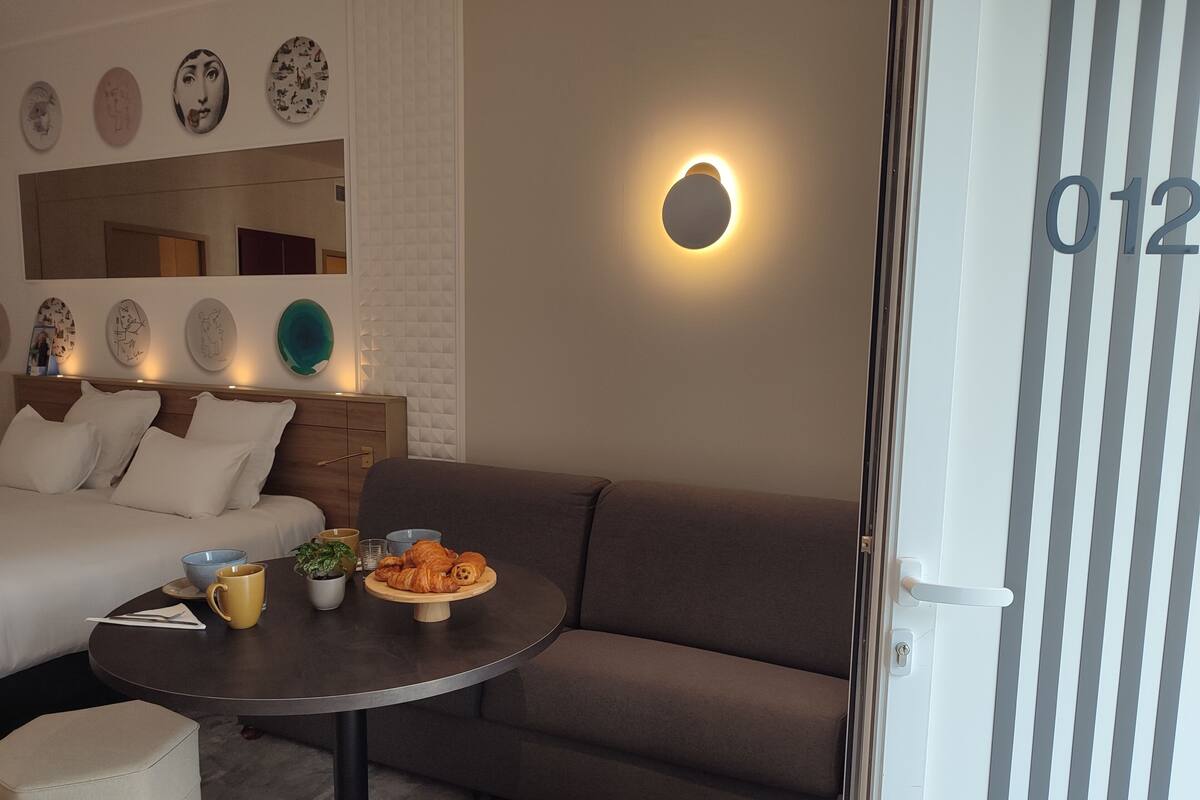 Un salon moderne avec un canapé, une table ronde sur laquelle se trouve une assiette de croissants, une tasse de café et un bol. Les murs sont décorés et il y a un miroir.
