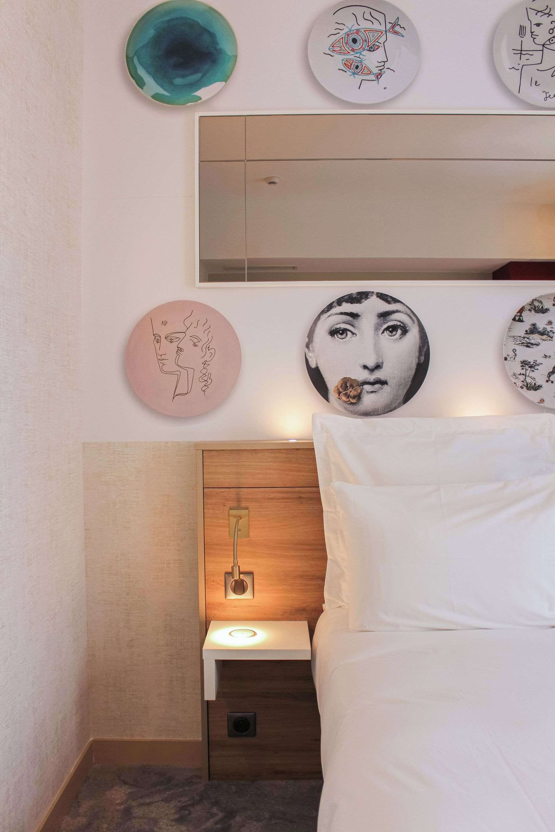 A hotel room with a bed, a nightstand with a lamp, and wall art including plates with designs.
