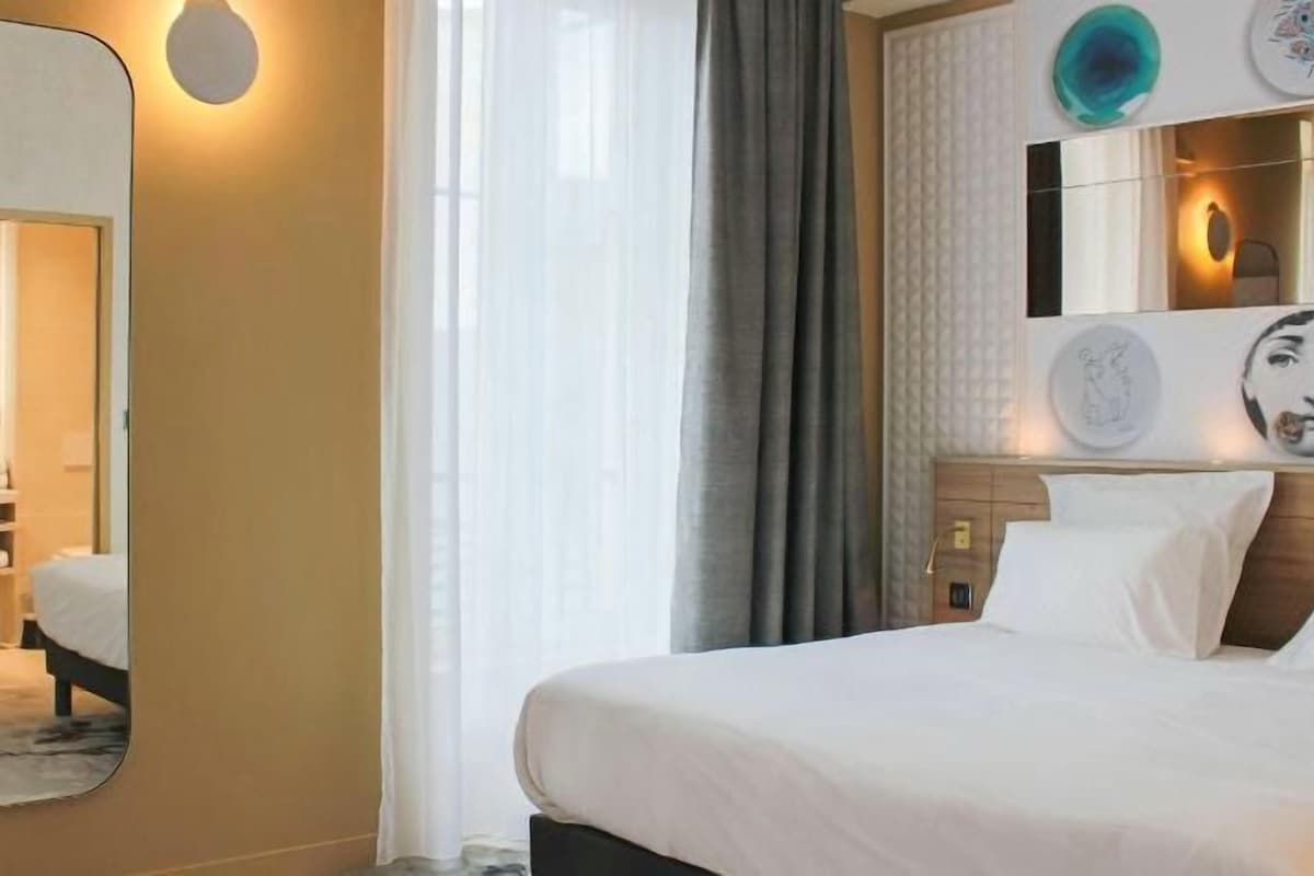 Hotellihuone, jossa on sänky, peili, yöpöytä ja seinään kiinnitetty valaisin.