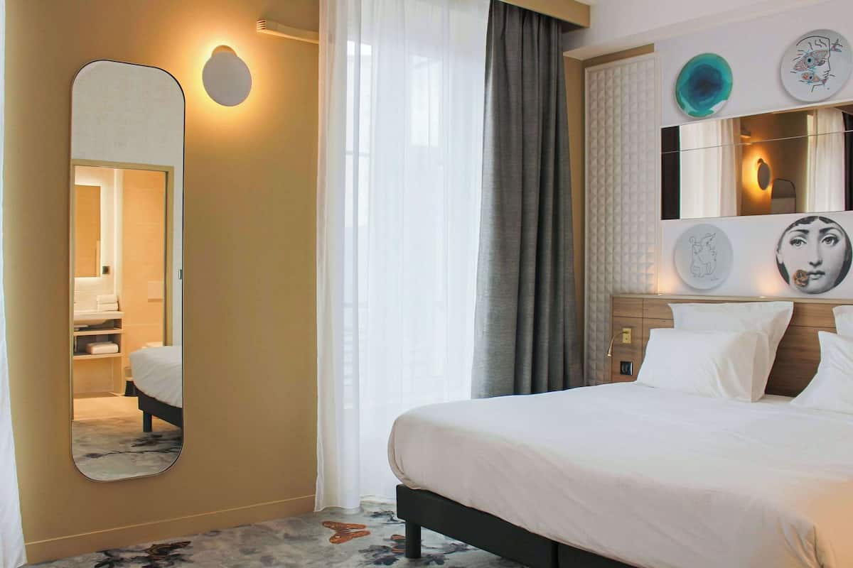 Une chambre d’hôtel comprenant un lit, un grand miroir, une table de chevet et une œuvre d’art accrochée au mur.