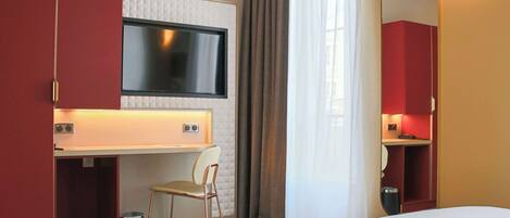 Quarto de hotel com uma cama, uma mesa com cadeira, televisão e espelho.