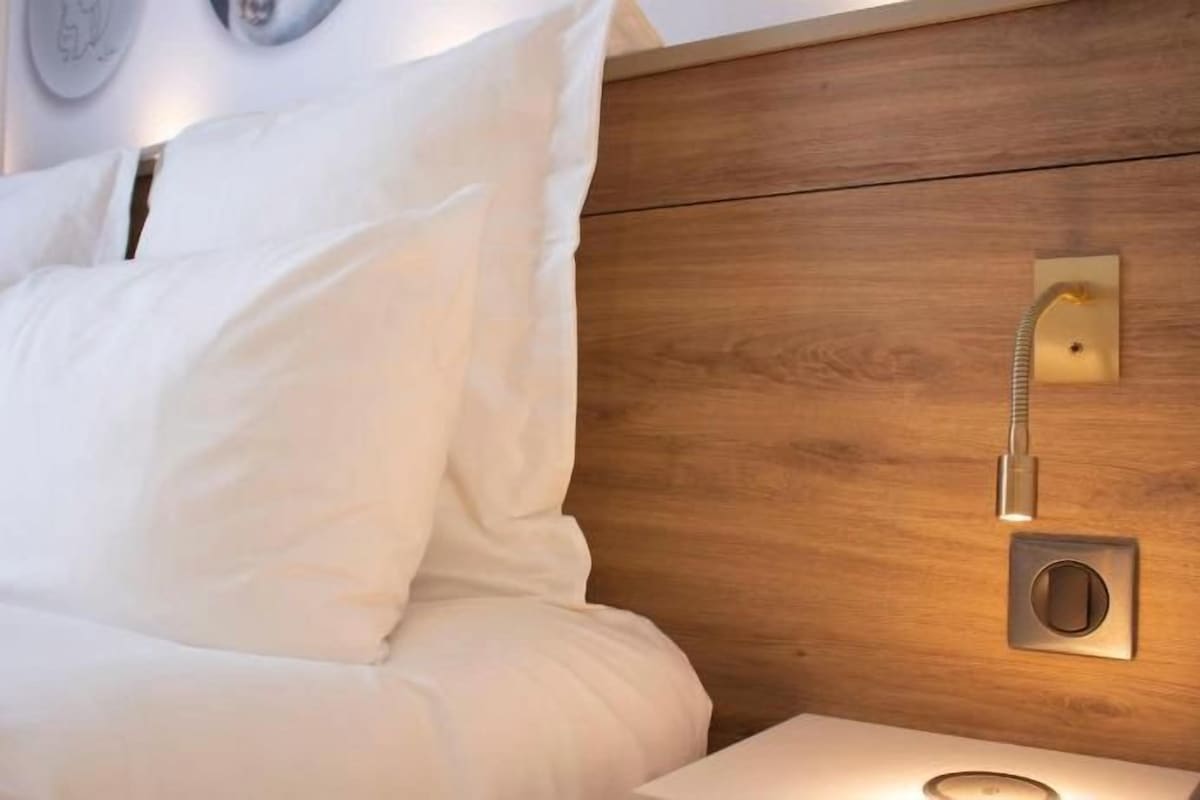 Una cama bien tendida con sábanas blancas, cabecera de madera y mesita de noche con lámpara.
