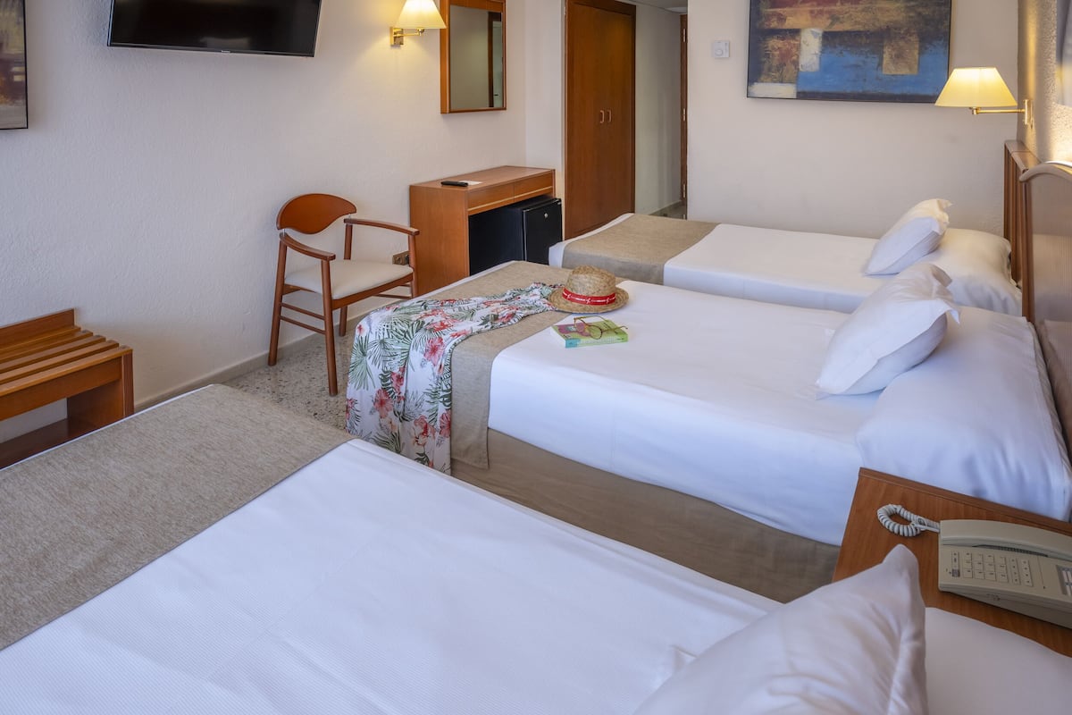 Quarto de hotel com três camas de solteiro, uma escrivaninha com computador, uma cadeira e uma televisão pequena.