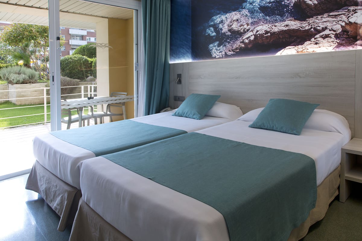 Quarto de hotel com duas camas, cada uma com colcha e almofadas verdes combinando. Há uma grande janela com vista para um jardim e edifícios lá fora.