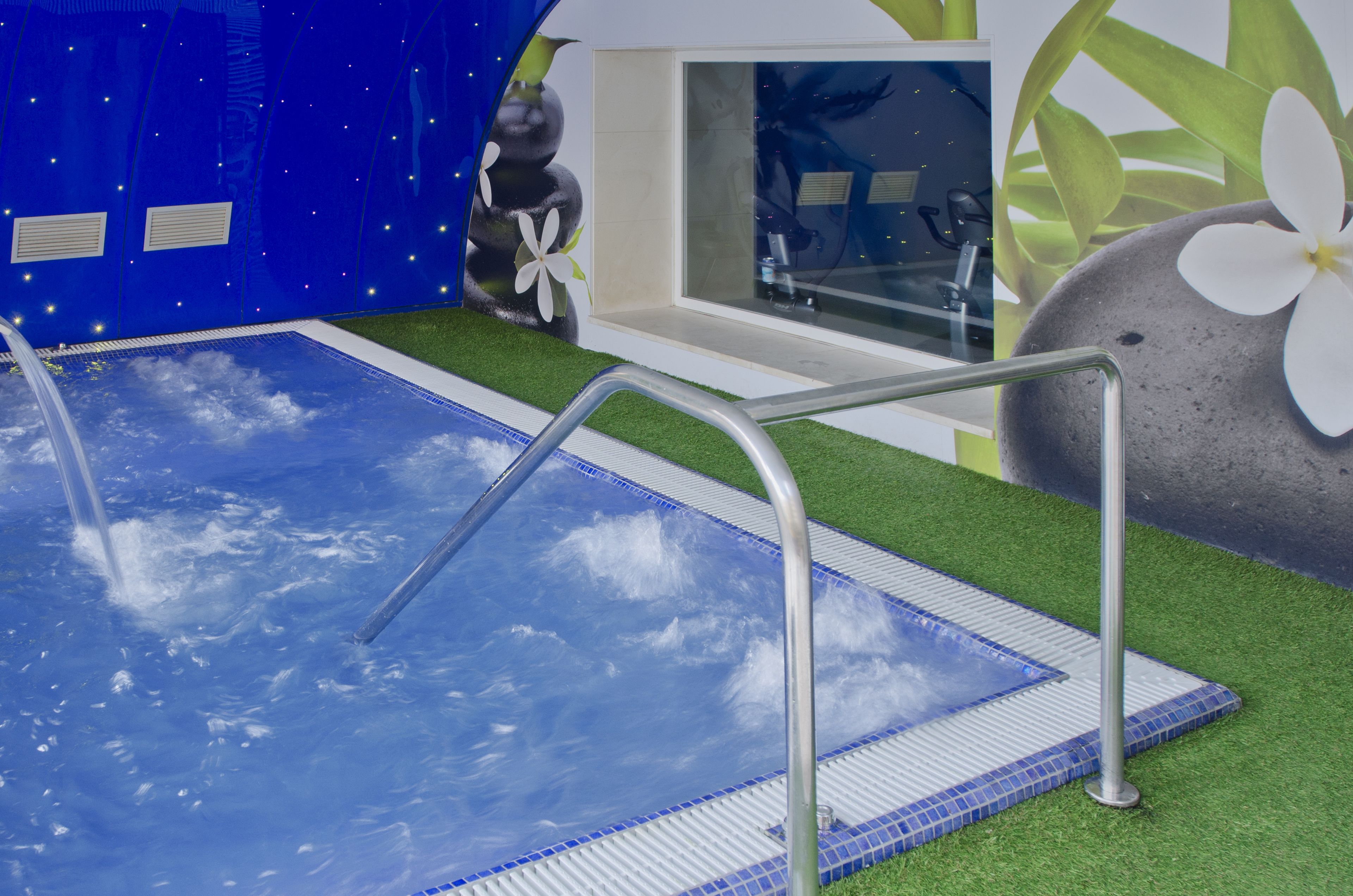 A swimming pool with a glass bottom, surrounded by artificial grass and a stone feature with a white flower.
