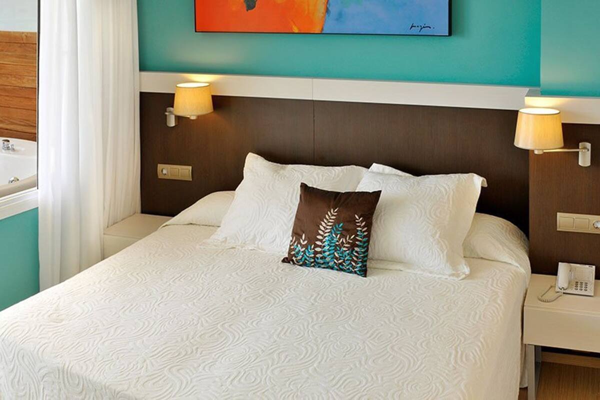 Quarto de hotel com cama, duas luminárias de cabeceira, mesa de cabeceira com telefone, banheiro com pia e espelho, e um quadro na parede.