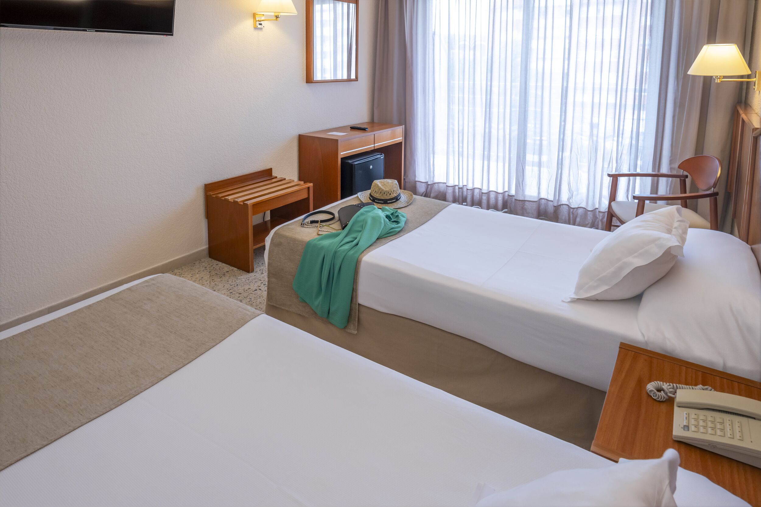 Quarto de hotel com duas camas, uma escrivaninha, uma cadeira, uma mesinha, uma lâmpada, uma televisão e uma janela com cortinas.