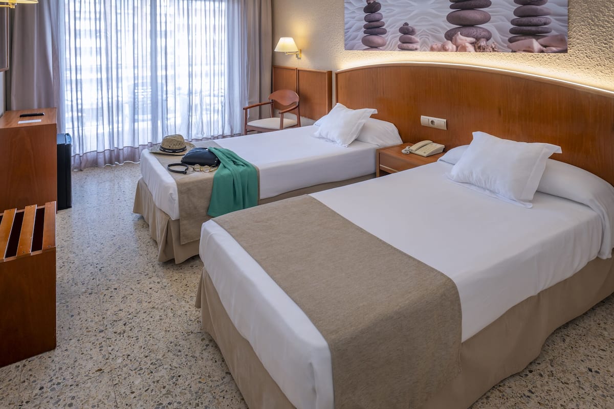 Quarto de hotel com duas camas, uma escrivaninha, uma cadeira, um telefone e uma luminária fixada na parede.