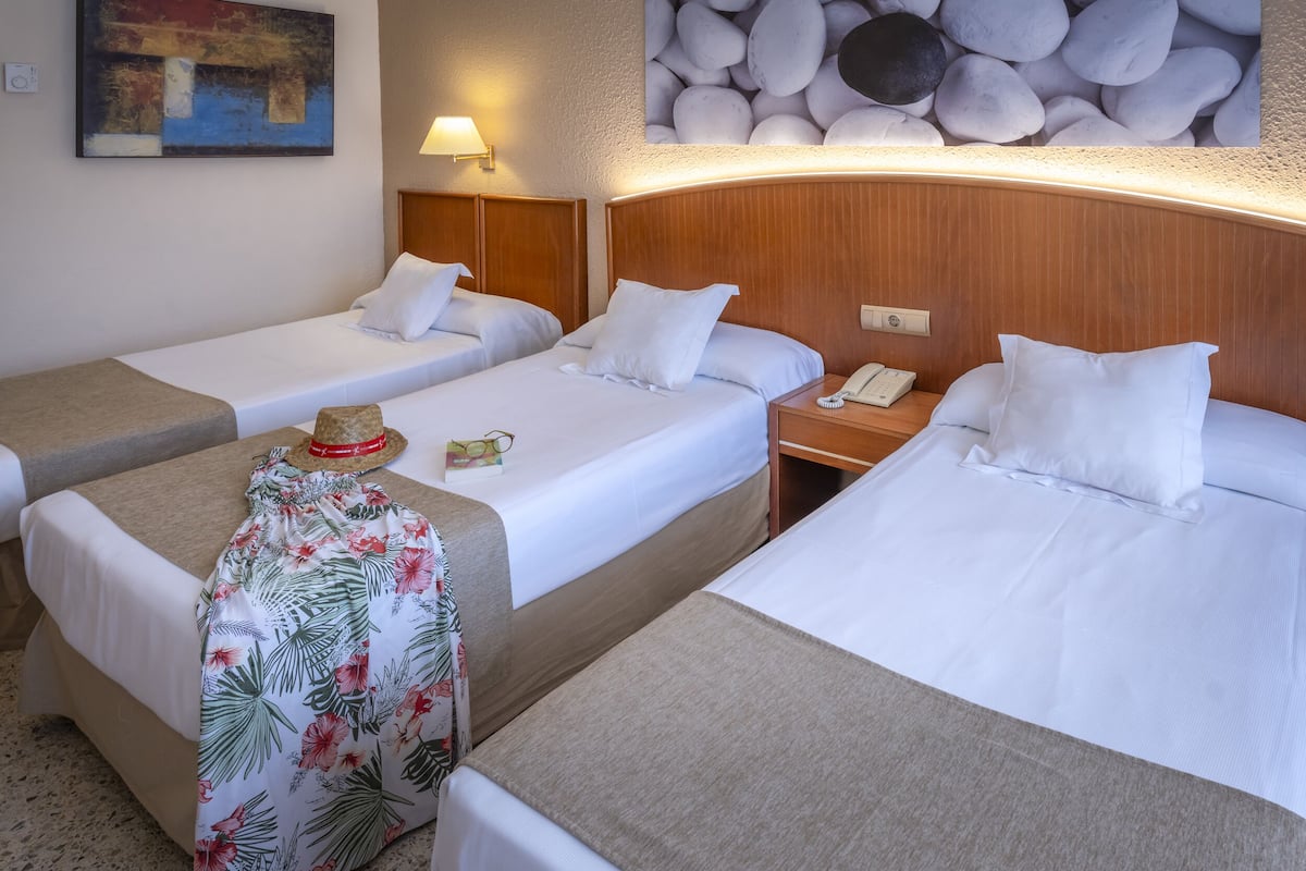 Quarto de hotel com duas camas de solteiro, uma pequena mesa com um chapéu e uma caneca, uma luminária e um telefone na mesa de cabeceira.