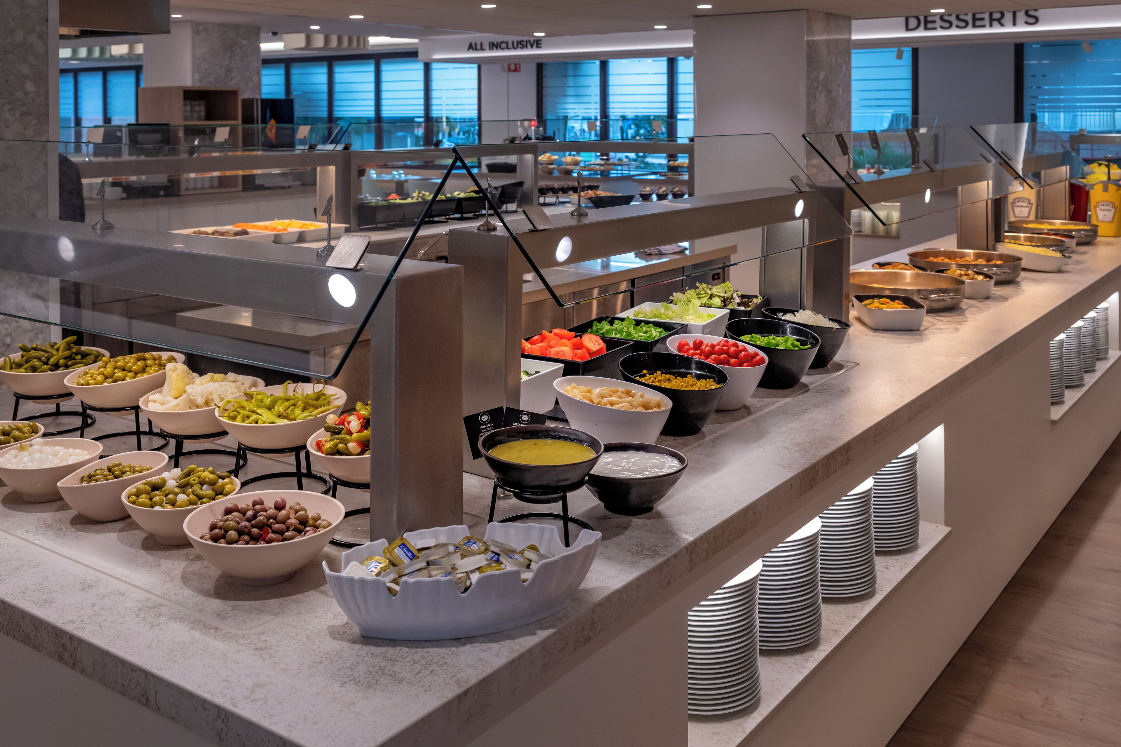Zona de bufé con diversas opciones de comida, como ensaladas, salsas y postres, dispuestas sobre una larga mesa con una mampara de vidrio.