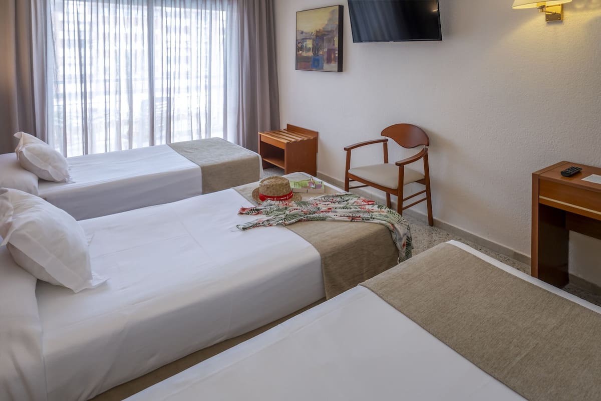 Quarto de hotel com duas camas de solteiro, uma cadeira de madeira, uma mesa pequena, televisão fixada na parede e uma janela com cortinas.