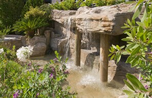 Une cascade d’eau se déversant sur une formation rocheuse, le tout étant entouré d’une végétation luxuriante et de plantes en fleurs.
