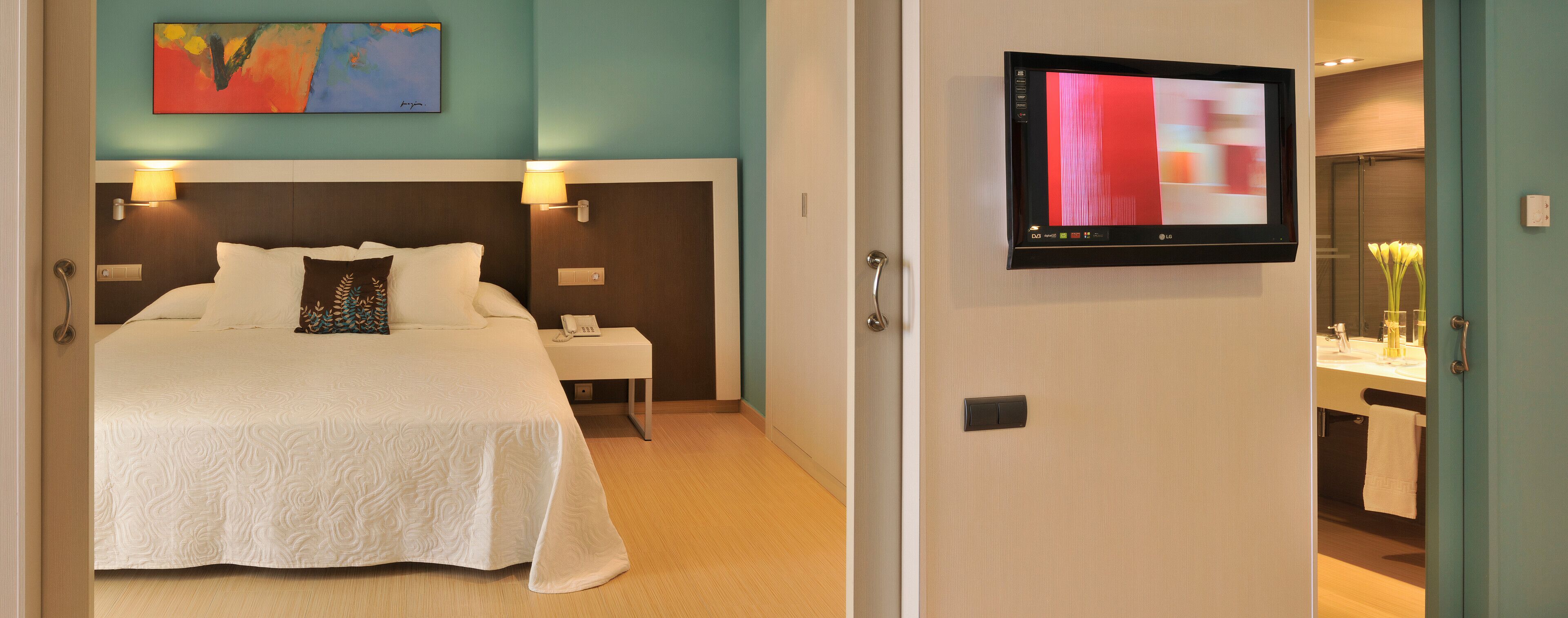 Habitación de hotel con cama, lámparas de noche, televisión y baño visible a través de una puerta abierta.