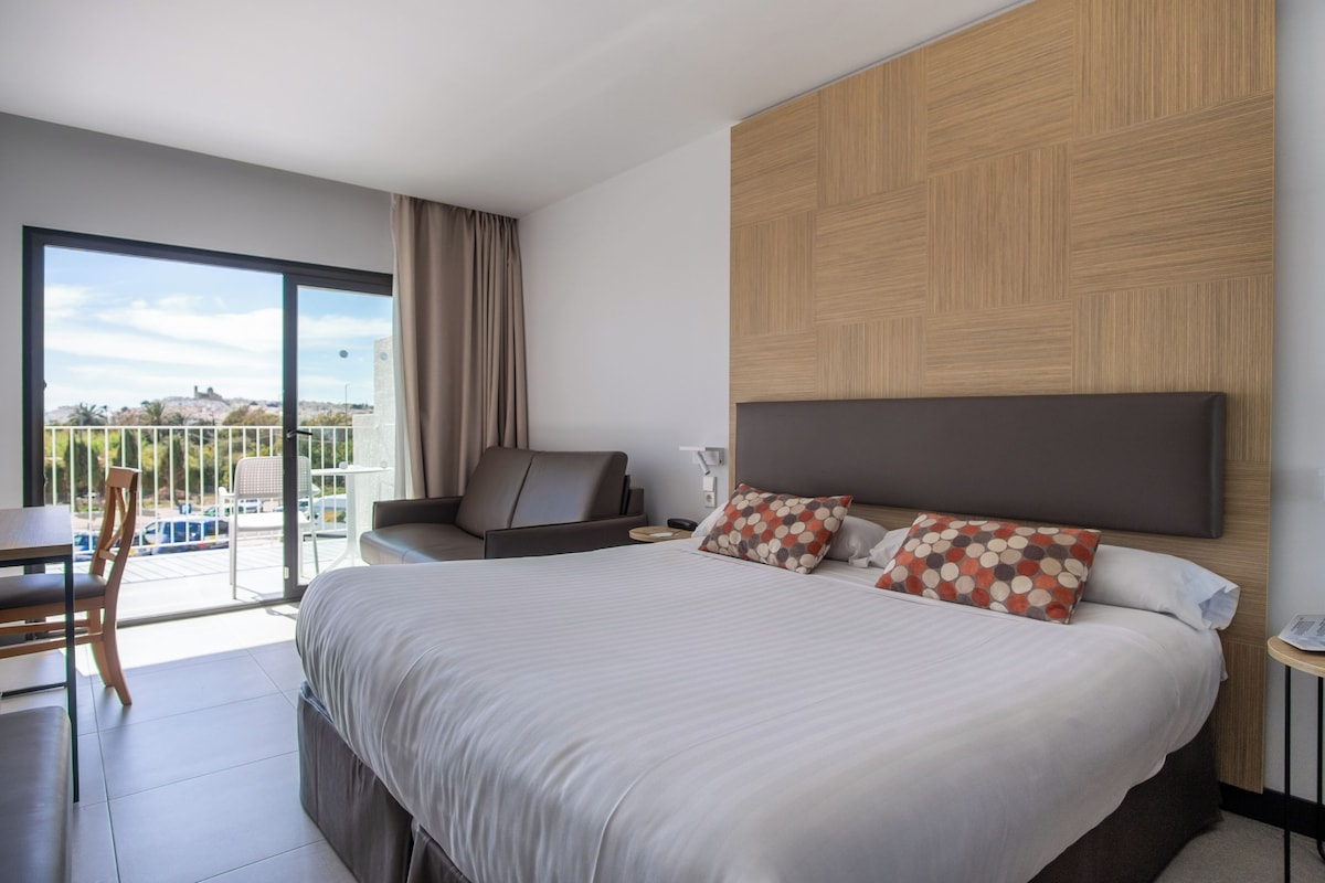 Une chambre d’hôtel moderne dotée d’un grand lit, d’un coin salon avec une chaise et une table, et d’un balcon offrant une vue agréable.