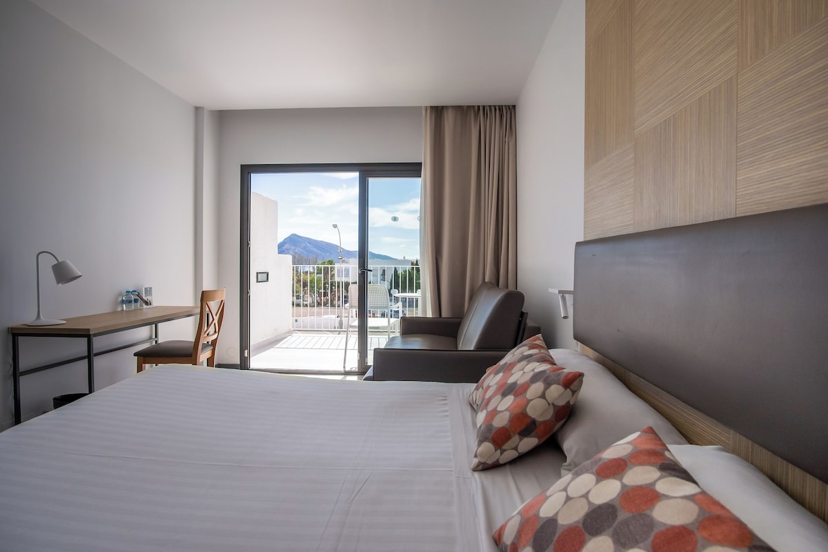 Une chambre d’hôtel avec un lit, un bureau, une chaise et un balcon offrant une vue sur les montagnes.