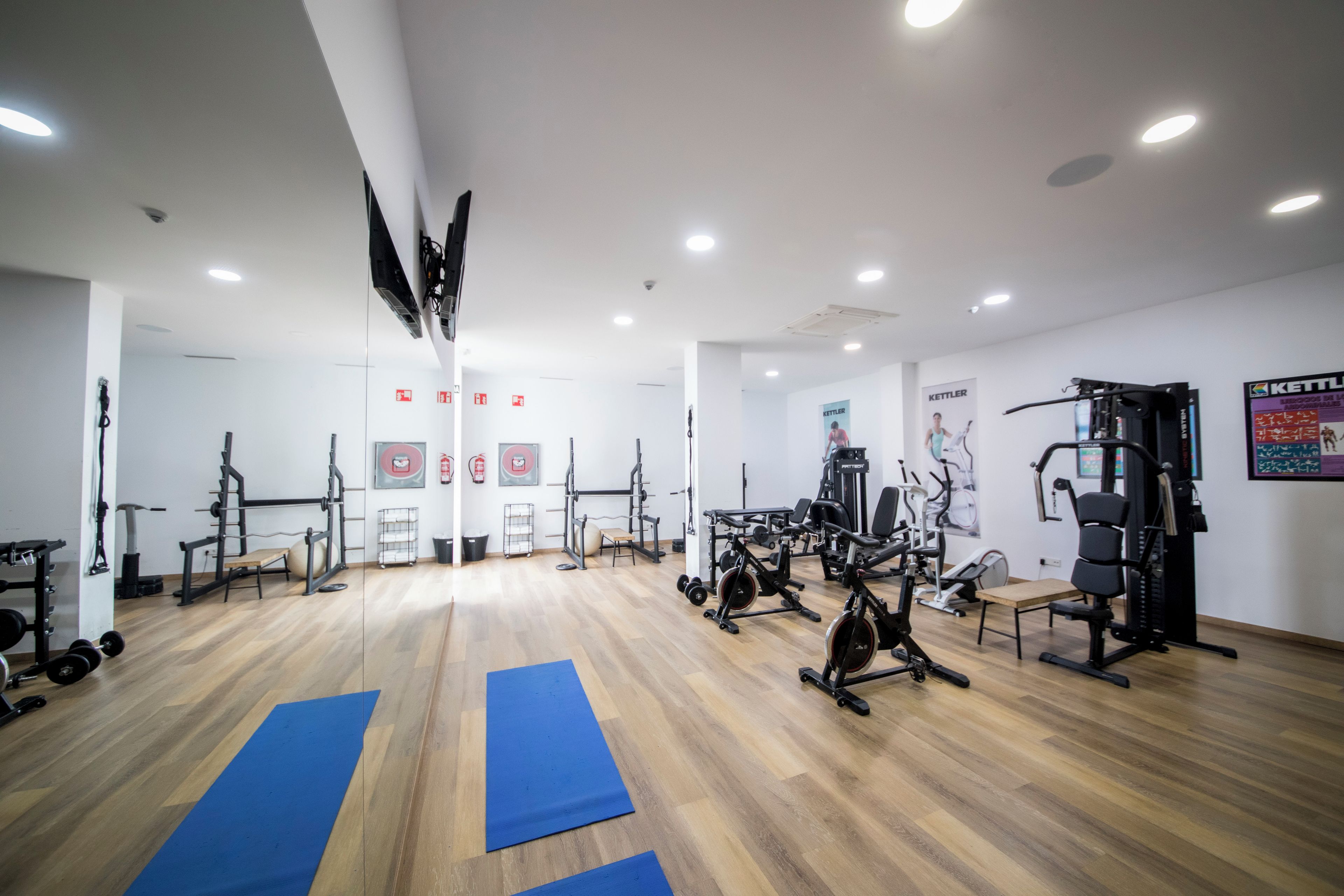 Et fitnesscenter med forskellig træningsudstyr, blå måtter på gulvet og et hvidt loft med indbyggede spots.