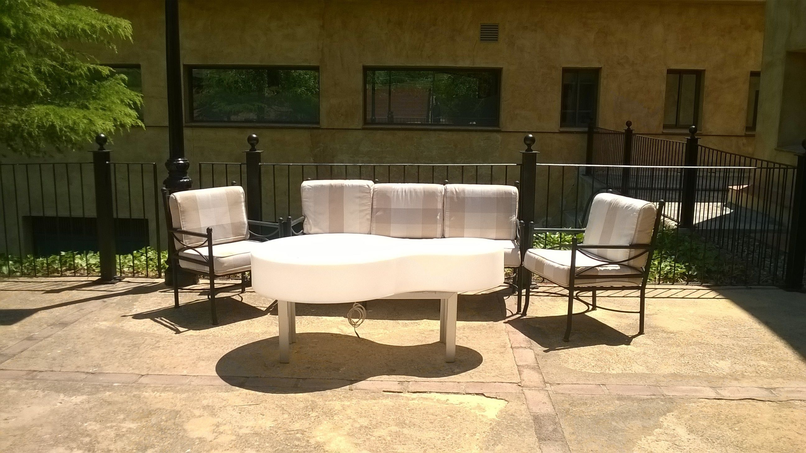 An outdoor patio with a round table and four chairs, a metal fence, and a building with windows in the background.